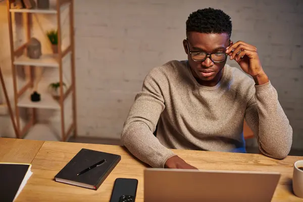 Серйозний афроамериканський фрілансер в окулярах думає біля ноутбука, що працює з дому вночі — стокове фото