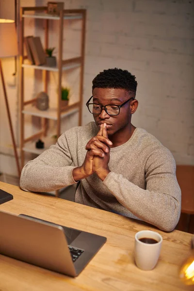 Jovem freelancer afro-americano em óculos pensando perto de laptop trabalhando de casa à noite — Fotografia de Stock
