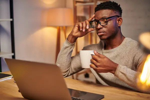 Reflexivo freelancer afroamericano con taza de café sentado cerca de la computadora portátil en la oficina en casa - foto de stock