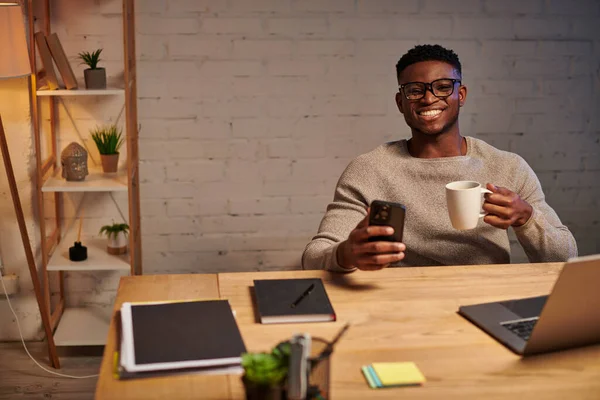 Joyeux freelance afro-américain avec café et smartphone près d'un ordinateur portable la nuit au bureau à domicile — Photo de stock