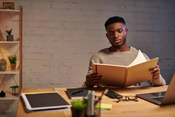 Joven freelancer afroamericano reflexivo mirando el cuaderno mientras trabaja en casa por la noche - foto de stock