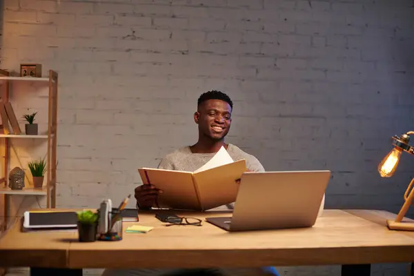 Allegro freelance afro-americano con notebook che guarda il portatile mentre lavora a casa di notte — Foto stock