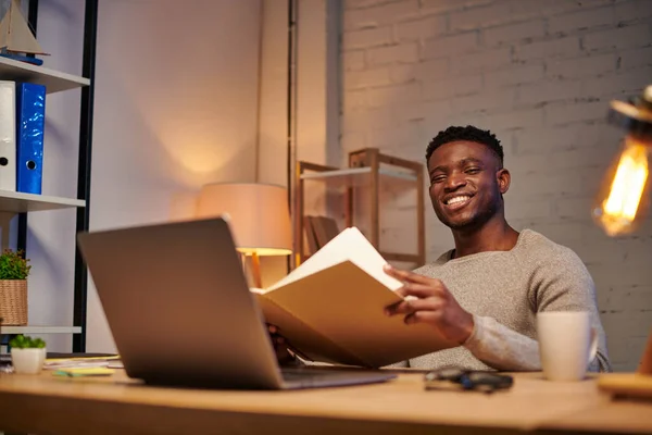 Feliz afroamericano freelancer con portátil mirando portátil mientras trabaja en casa por la noche - foto de stock