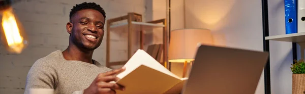 Piacere afro-americano freelance con notebook guardando computer portatile in home office di notte, banner — Foto stock