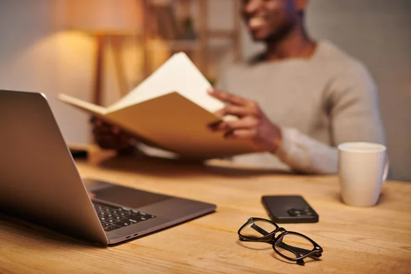 Focus sur les lunettes et l'ordinateur portable avec smartphone près de l'homme afro-américain cultivé dans le bureau à domicile — Photo de stock