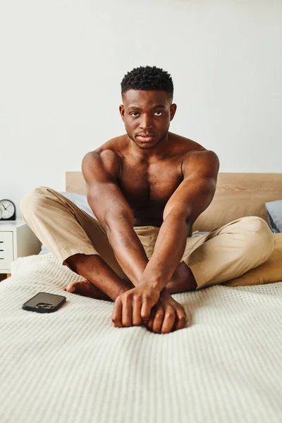 Selbstbewusster und starker afrikanisch-amerikanischer Mann in Pyjamahosen, der auf dem Bett sitzt und in die Kamera schaut — Stockfoto