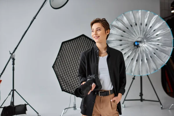 Jolly female photographer in everyday clothes with camera in hands smiling and looking away — Stock Photo