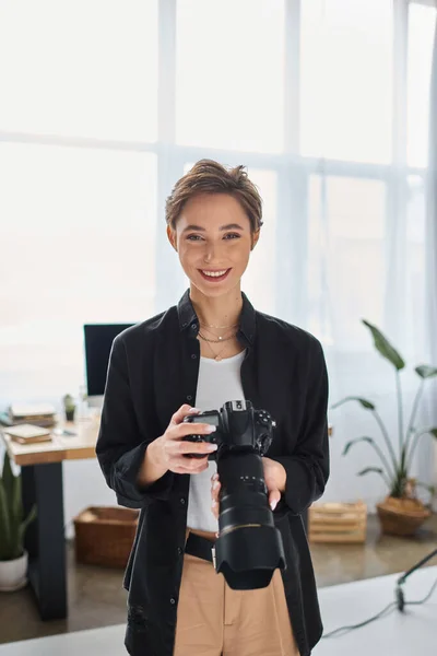 Joyeuse femme photographe aux cheveux courts en tenue décontractée posant avec caméra et souriant joyeusement — Photo de stock