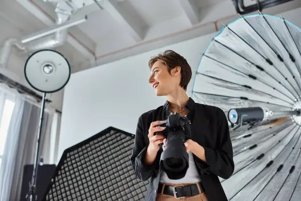 Fröhliche Fotografin in lässiger Kleidung mit Kamera in den Händen, lächelnd und wegschauend — Stockfoto