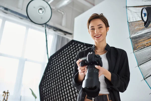 Joyous young woman with short hair in everyday clothes posing in her studio with camera in hands — Stock Photo