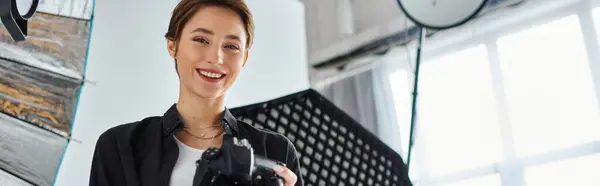 Femme joyeuse avec les cheveux courts dans les vêtements de tous les jours posant dans son studio avec caméra dans les mains, bannière — Photo de stock