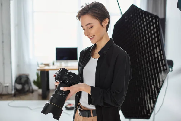 Merry female photographer in casual outfit with camera in her hands smiling and looking at photos — Stock Photo