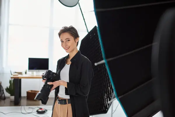 Jolly pretty woman with short hair in everyday clothes posing in her studio with camera in hands — Stock Photo