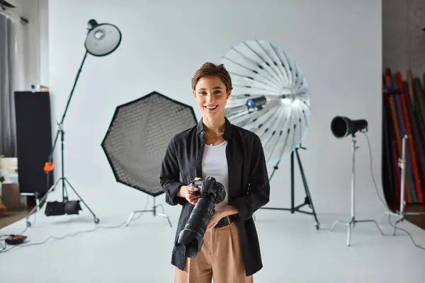 Happy pretty woman with short hair in everyday clothes posing in her studio with camera in hands — Stock Photo