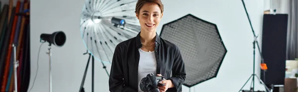 Joyful pretty woman with short hair in everyday clothes posing in her studio with camera in hands — Stock Photo