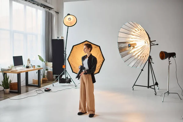 Attrayant jeune photographe femme en tenue quotidienne posant dans son studio et détournant les yeux — Photo de stock