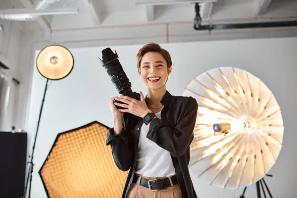 Cheerful pretty woman with short hair in everyday clothes posing in her photo studio with camera — Stock Photo