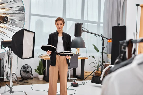 Mulher atraente alegre com cabelo curto posando com seu equipamento de fotografia em seu estúdio — Fotografia de Stock