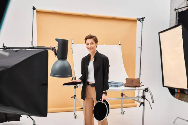 Mujer alegre de pelo corto con equipo de fotografía sonriendo alegremente a la cámara en su estudio - foto de stock