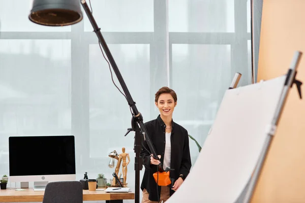 Beautiful joyous woman with photography equipment smiling cheerfully at camera in her studio — Stock Photo