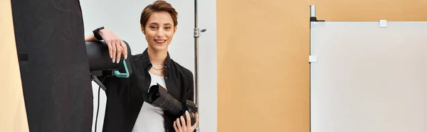 Mujer atractiva feliz con equipo de fotografía sonriendo felizmente a la cámara en su estudio, pancarta - foto de stock