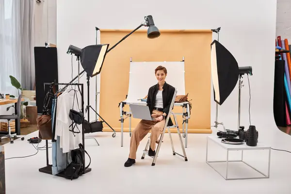 Cheerful pretty woman sitting with laptop while retouching photos and smiling at camera in studio — Stock Photo