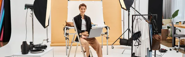 Mujer alegre sentado con el ordenador portátil mientras retocando fotos y sonriendo a la cámara en el estudio, pancarta - foto de stock