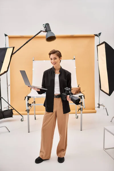 Happy attractive woman smiling joyfully while retouching photos and holding her camera in studio — Stock Photo