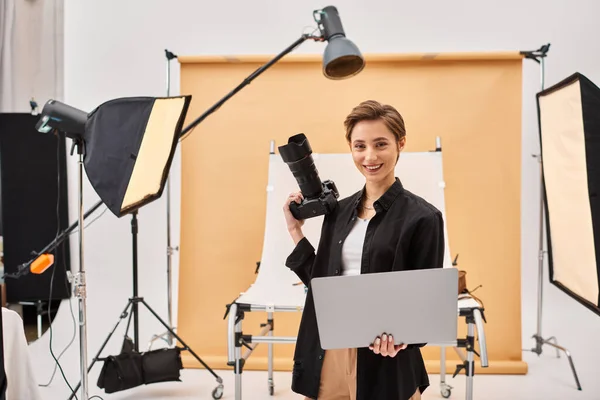 Cheerful attractive woman smiling happily while retouching photos and holding her camera in studio — Stock Photo