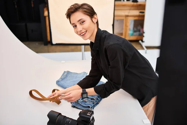 Joyful female photographer preparing to make photos of jeans and belt and smiling at camera — Stock Photo
