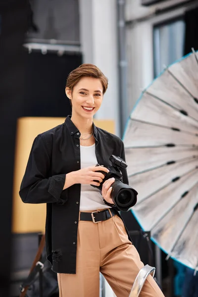 Happy beautiful woman in casual attire smiling at camera and holding camera in her photo studio — Stock Photo