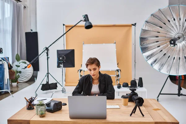 Giovane bella fotografa donna in abbigliamento casual che lavora al suo computer portatile con una tazza di caffè sul tavolo — Foto stock
