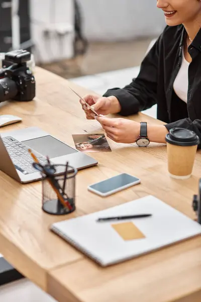Cropped view of laptop and camera on table next to cheerful female photographer with photos in hands — Stock Photo