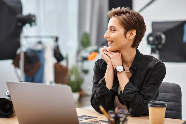 Young cheerful woman in casual attire sitting at desk in her photo studio and looking away — Stock Photo