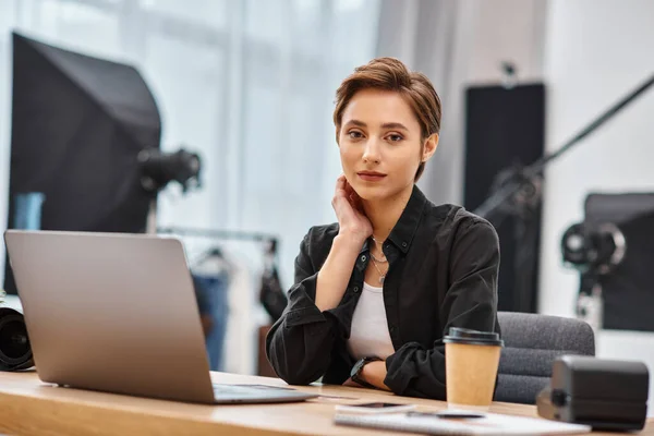 Young beautiful woman in casual attire sitting at desk in her photo studio and looking at camera — Stock Photo