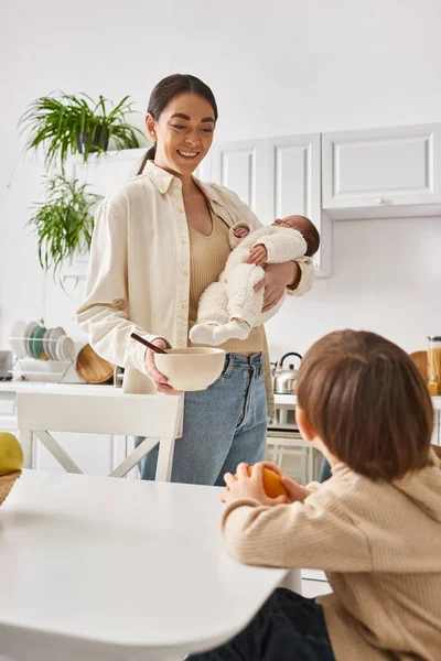 Cheerful mother in casual attire preparing breakfast for her toddler son while holding her newborn — Stock Photo