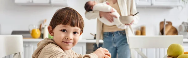 Jolly mother in casual attire preparing breakfast for her toddler son while holding newborn, banner — Stock Photo