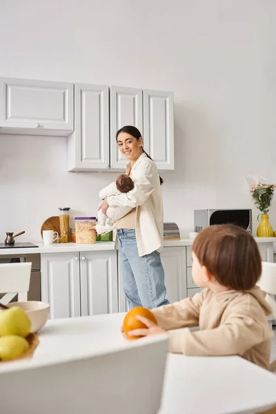 Happy mother in casual attire preparing breakfast for her toddler son while holding her newborn — Stock Photo