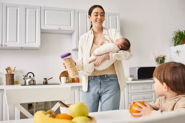 Happy mother in casual attire holding cornflakes near her toddler son while holding her newborn — Stock Photo