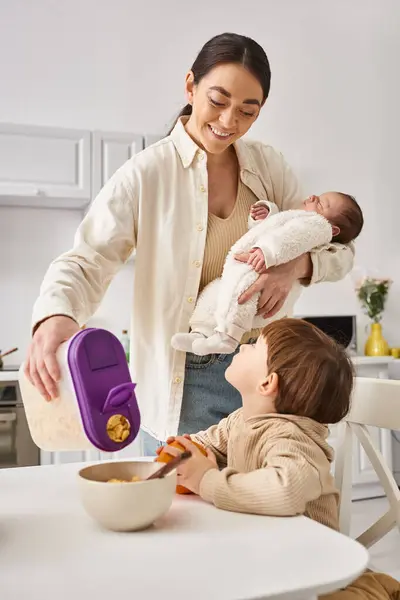Merry woman pouring delicious corn flakes to her adorable toddler son while holding her newborn baby — Stock Photo