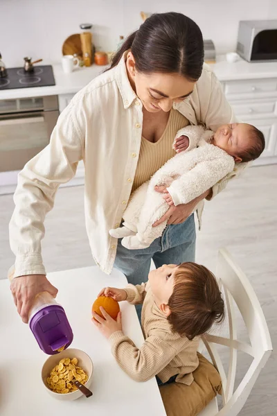 Merry beautiful woman pouring corn flakes to her adorable toddler son while holding her newborn baby — Stock Photo
