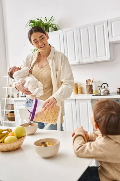 Mulher bonita feliz derramando flocos de milho para seu filho adorável criança enquanto segurando seu bebê recém-nascido — Fotografia de Stock