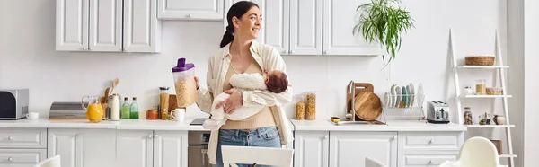 Beautiful joyous woman in comfy homewear holding her cute newborn baby while preparing breakfast — Stock Photo
