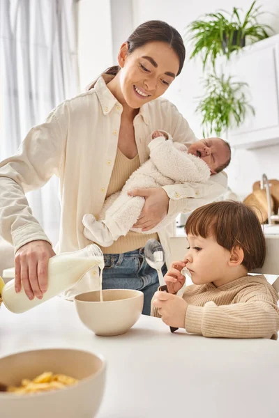 Cheerful woman pouring milk into corn flakes for her toddler son and holding her newborn baby — Stock Photo