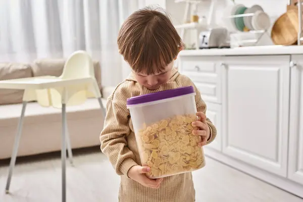 Adorable toddler boy in casual homewear holding pack of corn flakes during breakfast on kitchen — Stock Photo