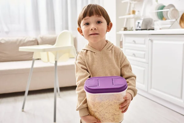 Adorable little boy in casual homewear holding pack of corn flakes during breakfast on kitchen — Stock Photo