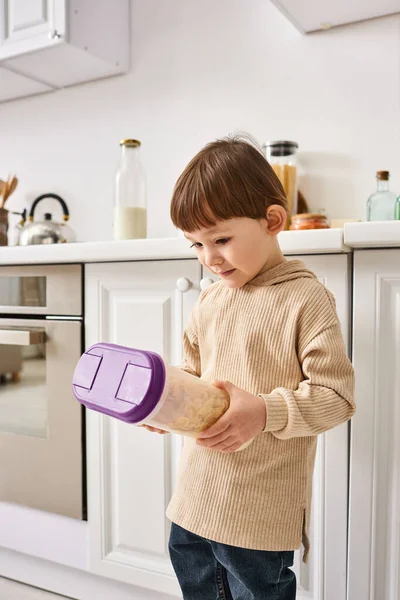 Adorable toddler boy in cozy homewear holding pack of corn flakes during breakfast on kitchen — Stock Photo