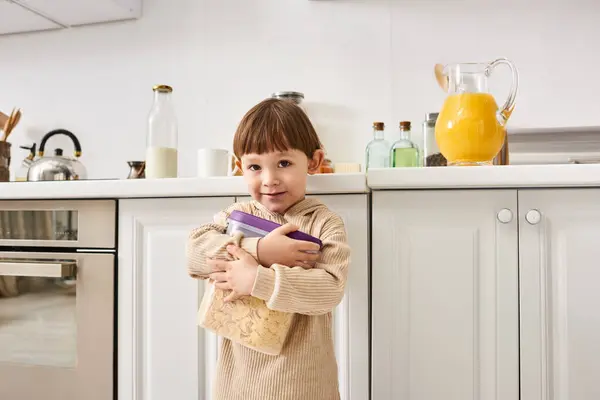 Adorable little boy in comfortable homewear holding pack of corn flakes during breakfast on kitchen — Stock Photo