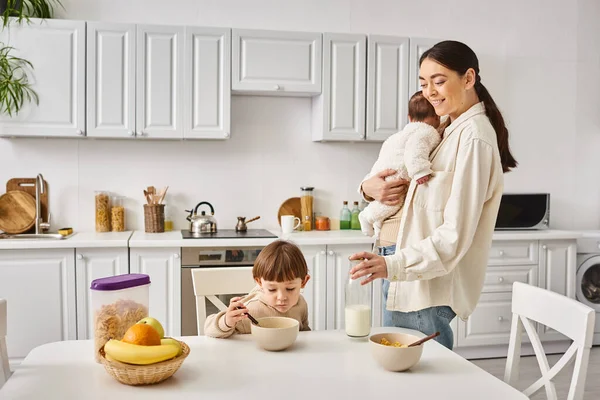 Cheerful beautiful mother posing next to her toddler son with newborn in hands during breakfast — Stock Photo