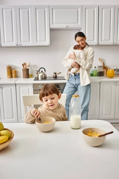Joyous loving mother posing next to her toddler son with newborn in hands during breakfast — Stock Photo